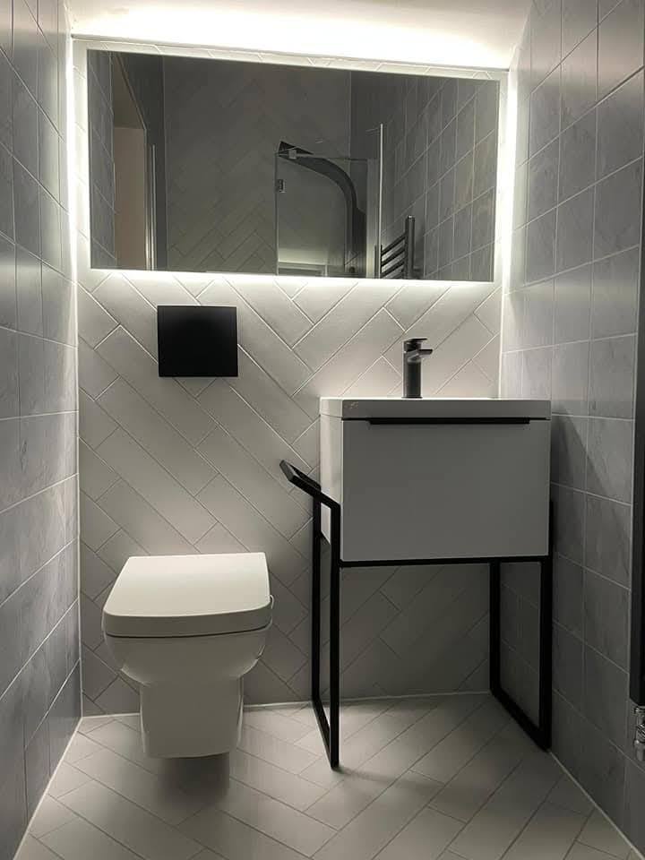 Finished cloakroom with grey herringbone tile, backlit mirror, wall-hung WC and small vanity with black frame legs